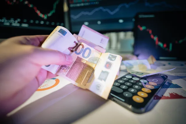 A hand holds euro notes with financial charts and a calculator, symbolizing investment and trading. Kosten Nachfolgeberatung: Honorare und Umsatzsteuer